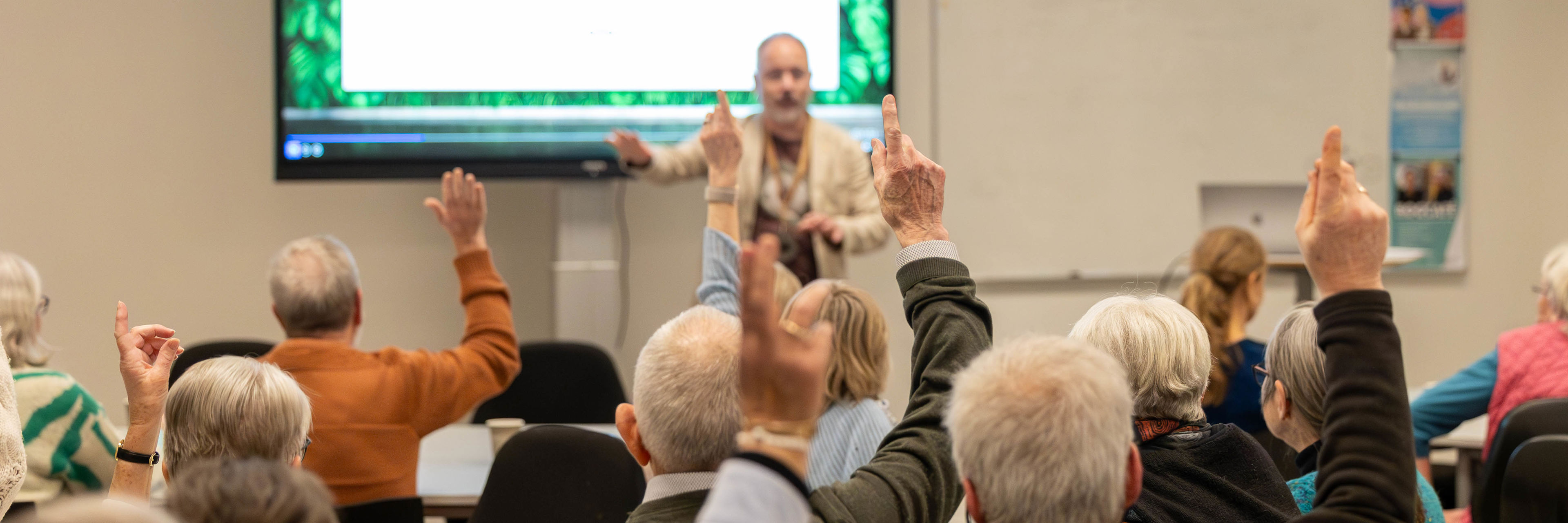 Møde. En mand står med fronten mod kamera og deltagere sidder med ryggen til og rækker hånden op